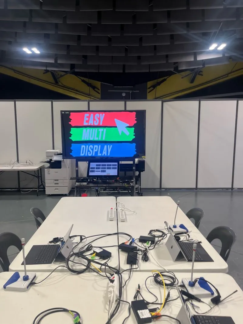 Sala di controllo temporanea di Lourdes 2026: due portatili e microfoni sul tavolo, lo schermo touch che pilota la Multiprise HDMI e la schermata iniziale di Easy Multi Display