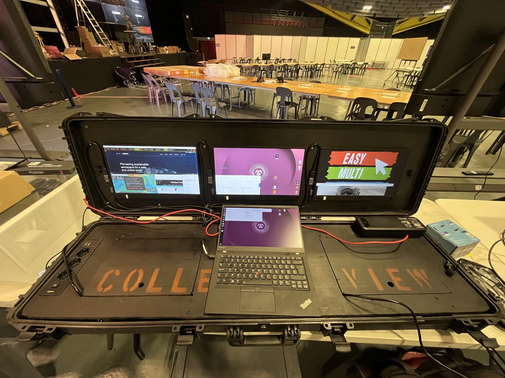 Valise de commandement EMD ouverte dans la salle de contrôle de Lourdes 2026, le logo Easy Multi Display sur l'écran, les tables des services en fond